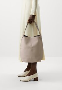 Růžová kožená kabelka s texturovaným povrchem, zaobleným tvarem a dlouhým ramenním popruhem. Na pozadí světle zbarvené šaty a pozadí.