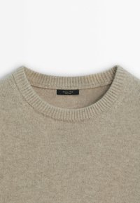 Pull beige à col rond avec col côtelé et texture, comportant une petite étiquette noire cousue à l'intérieur de l'encolure.