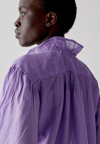 Blusa lavanda con colletto alto, schiena plissettata e tessuto morbido e leggero. Presenta una texture leggermente trasparente e una vestibilità rilassata.