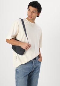 Hallhalli hall crossbody kott, millel on sile tekstuur ja kõver kuju, hoiab inimene, kes kannab heledat beežikat T-särki ja siniseid teksaseid.