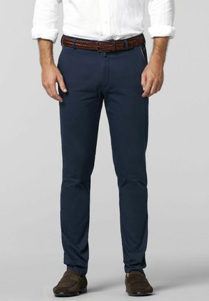 Homme portant une chemise blanche à manches longues rentrée dans un pantalon bleu marine avec une ceinture tressée marron et des mocassins en daim marron, debout devant un fond gris.