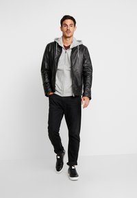 Zwarte leren jas over een grijze hoodie, gecombineerd met een donkere jeans en zwarte sneakers. De jas heeft een ritssluiting en subtiele stikdetails.
