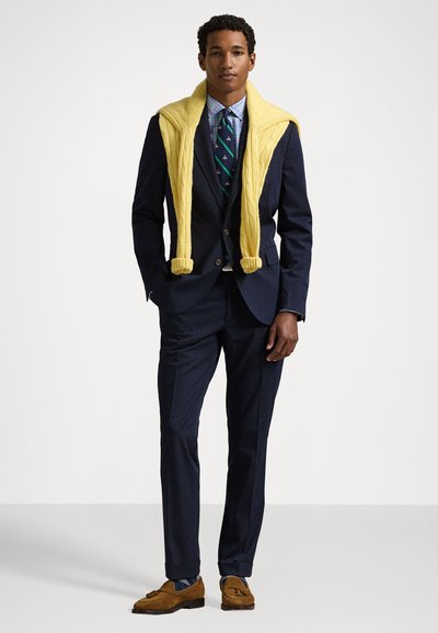 Costume navy avec un pull en tricot jaune clair drapé sur les épaules. Chemise à carreaux et cravate à motifs. Des mocassins marron complètent la tenue.