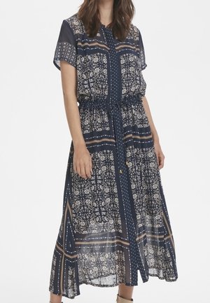 Femme portant une robe midi bleu marine à manches courtes avec des motifs floraux et géométriques blancs et beiges complexes.