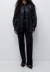 Chemise surdimensionnée en similicuir noir avec boutons-pression, col pointu et poche avant, associée à un jean flare noir et des bottines pointues.
