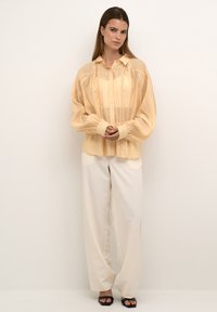 Hellgelbe Bluse mit aufwendigen Details und puffartigen Ärmeln, kombiniert mit lockeren cremefarbenen Hosen und schwarzen Sandalen. Einfaches, elegantes Design.