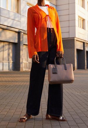 Personne portant un chemisier transparent orange, un haut blanc, un pantalon large noir, des sandales, tenant un sac à main beige avec des poignées noires dans un cadre urbain.