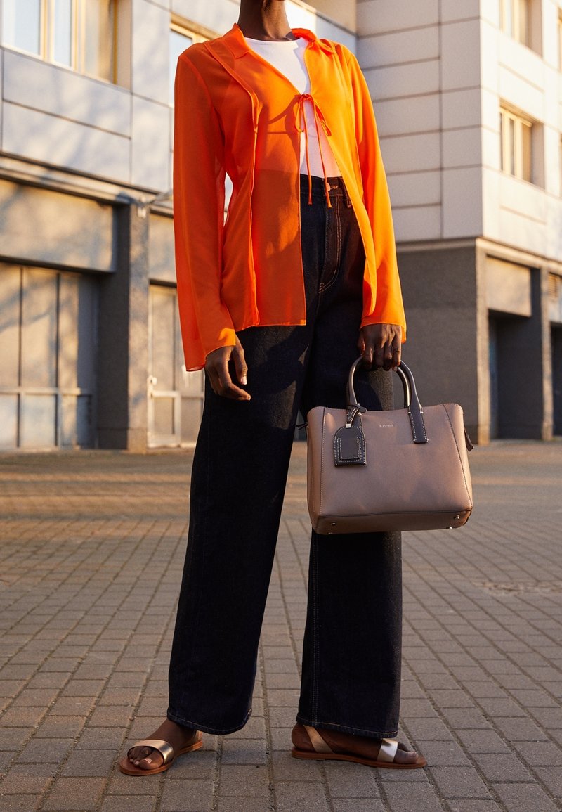 Personne portant un chemisier transparent orange, un haut blanc, un pantalon large noir, des sandales, tenant un sac à main beige avec des poignées noires dans un cadre urbain.
