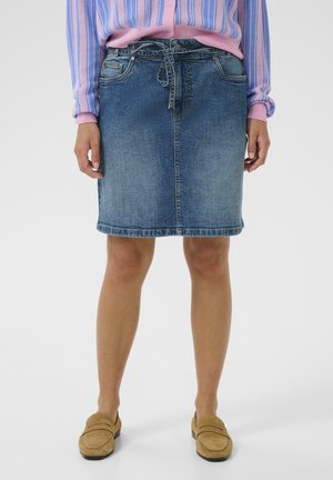 Frau trägt einen knielangen blauen Jeansrock mit gebundenem Gürtel, einen rosa-blau gestreiften Pullover und hellbraune Wildleder-Loafers, steht vor weißem Hintergrund.