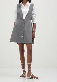Graues ärmelloses Kleid mit tiefem V-Ausschnitt, Knöpfen vorne und ausgestelltem Rock. Kombiniert mit silbernen Riemchensandalen auf nackten Beinen.