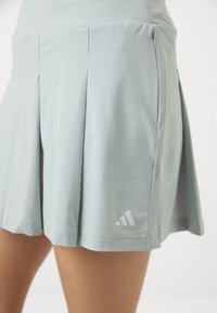 Világoskék sport szoknya, puha anyagból, pliszírozott fazonnal és egy apró fehér Adidas logóval az alján. Sima textúra és laza dizájn.