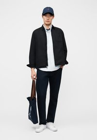 Νεαρός άνδρας με ναυτικό καπέλο, μαύρο μπουφάν, ριγέ πουκάμισο, σκούρα παντελόνια, λευκά αθλητικά παπούτσια, κρατώντας ναυτική τσάντα ώμου με καφέ χερούλια, στέκεται μπροστά σε λευκό φόντο.