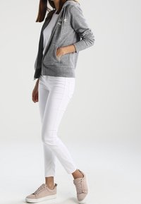 Šedá mikina na zip s přední kapsou a šňůrkou, nošená přes bílé tričko. Kombinováno s bílými kalhotami a světle růžovými teniskami.