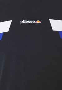 Czarny materiał z centralnie umieszczoną naszywką logo "ellesse", z półokrągłym wzorem w kolorze pomarańczowym i czerwonym, nad białymi i niebieskimi ukośnymi paskami.
