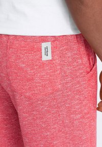 Un pantalon de jogging rouge chiné présente une texture douce, des poches latérales et une étiquette blanche avec "ombre menswear" sur la ceinture arrière.