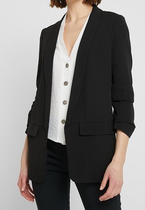 Vrouw draagt een zwarte, op maat gemaakte blazer met geplooide mouwen over een witte blouse met knopen en zwarte broek, staand tegen een effen achtergrond.
