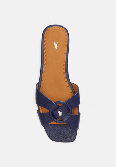 Sandalia de cuero azul marino con punta cuadrada y tiras cruzadas, con un adorno circular y plantilla marrón con relieve del logo.