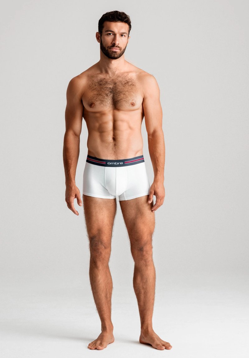 Vita boxershorts med en mörk midjeband med röda detaljer. Modellen står barfota och visar upp en åtsittande design och slät textur.