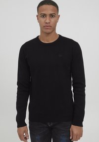 Czarny sweter z dzianiny, okrągły dekolt, slim fit, ściągacze na rękawach i dole, z delikatnym logo na piersi. Noszony ze ciemnymi dżinsami.