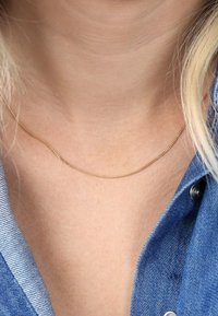 Dünne goldene Halskette liegt auf nackter Haut, mit einer zarten, glatten Textur und einem subtilen Schimmer, kombiniert mit einem Denimhemd.