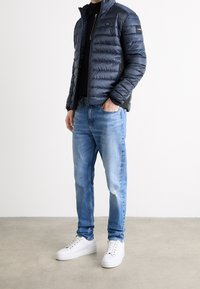 Marinblå puffjacka med kviltat mönster, svarta detaljer och dragkedjestängning, kombinerad med ljusblå jeans och vita sneakers.