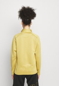 Osoba z kręconymi włosami w wysokim koku, ubrana w żółtą bluzkę z długim rękawem i czarne spodnie, stojąca na tle jednolitej białej ściany.