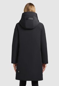 Sort polstret jakke med en stor hætte, der har et logo-patch på ryggen. Lange ærmer, lige snit og glat tekstur.