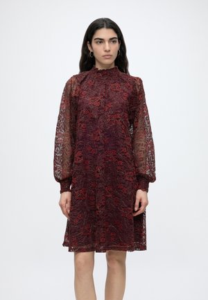 Γυναίκα που φοράει μια μπορντό δαντελένια φορέματα με floral μοτίβα, ψηλό βολάν στο λαιμό και μακριά διαφανή φουσκωτά μανίκια, στέκεται μπροστά σε ένα απλό φόντο.