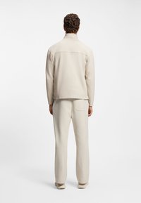 Beżowa kurtka na zamek z wysokim kołnierzem, zestawiona z dopasowanymi, zwężanymi spodniami. Miękki, lekki materiał, minimalistyczny design, z tylną kieszenią.