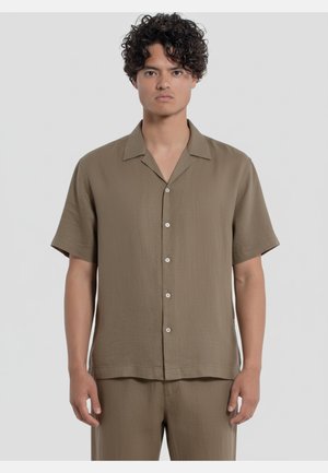 Uomo con capelli ricci che indossa una camicia marrone a maniche corte con bottoni e pantaloni abbinati su uno sfondo grigio uniforme.