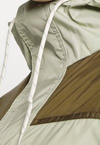 Primer plano de una chaqueta con capucha en verde claro y oliva, con cordones blancos y detalles de costura visibles.