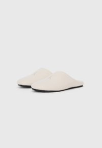 Witte wollen slippers met een gladde textuur, ronde neus, lage achterkant, en zwarte rubberen zool. Geborduurde letter "K" aan de voorkant.