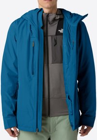 Uomo che indossa una giacca impermeabile blu aperta, sopra un fleece grigio della North Face con zip e pantaloni verde chiaro.