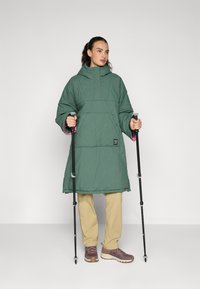 Poncho outdoor acolchado verde con un alto cuello, puños en rosa y un bolsillo frontal. Lleva puesto sobre pantalones beige y botas de senderismo marrones.