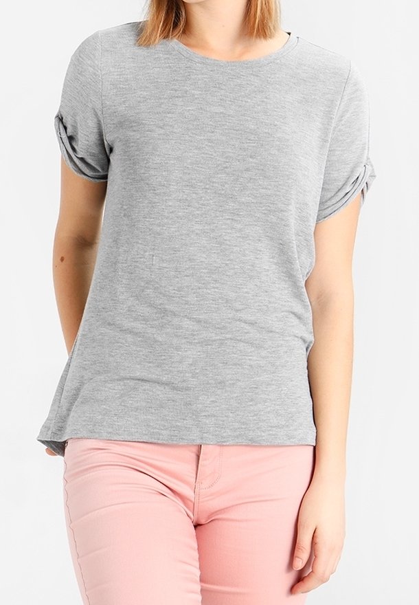 Femme portant un haut gris clair à manches courtes retroussées et un pantalon rose clair, debout devant un fond blanc.