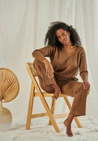 Brauner Strickpullover und passende Hose aus weichem Stoff. Das Model sitzt barfuß auf einem Holstuhl, mit einem dekorativen Fächer in der Nähe.
