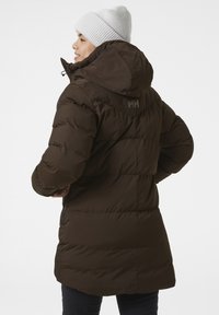 Bruine pufferjas met een capuchon, voorzien van geborduurde secties, ritszakken en een logo op de rug. Gebreid wollen muts in lichtgrijs.