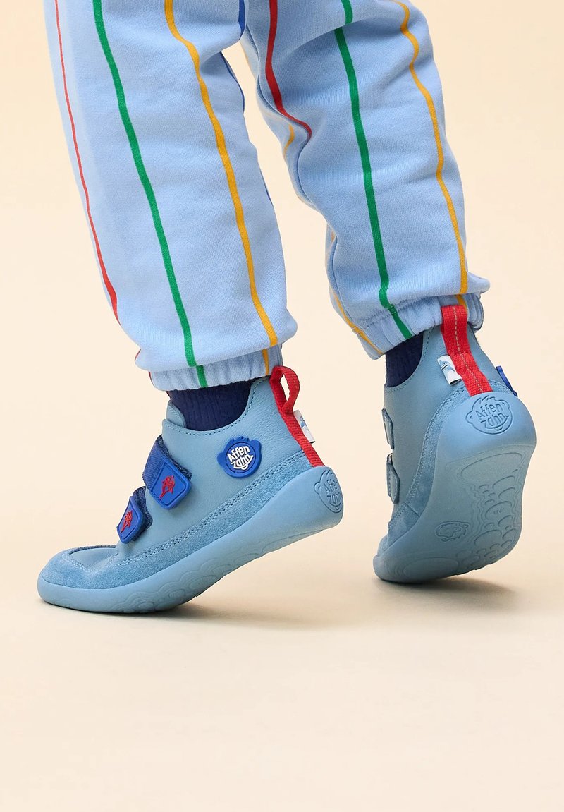Scarpe da bambino blu con cinturini in velcro, soletta in gomma, accenti rossi e blu e motivi testurizzati, abbinate a pantaloni a righe blu chiaro.
