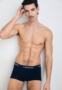 Navy blue Lacoste boxer shorts with a branded waistband. The fabric appears smooth, stretchable, and fitted to the model's waist.