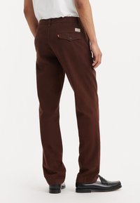Pantalones de algodón marrón con corte recto, bolsillo trasero con botón, y combinados con mocasines de cuero negros.