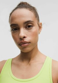 Nahaufnahme einer Person, die ein geripptes, leuchtend limettengrünes Tanktop trägt. Hautton ist hell mit natürlichem Make-up, das kleine goldene Ohrringe umfasst.