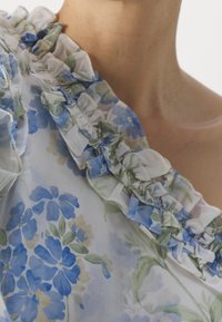 Blouse florale avec motifs bleus et verts, col froncé et tissu léger, conçue avec une silhouette décolletée.
