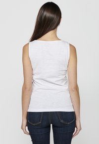 Top bianco senza maniche realizzato in cotone, con scollatura a barca e una texture liscia, abbinato a jeans aderenti blu scuro.