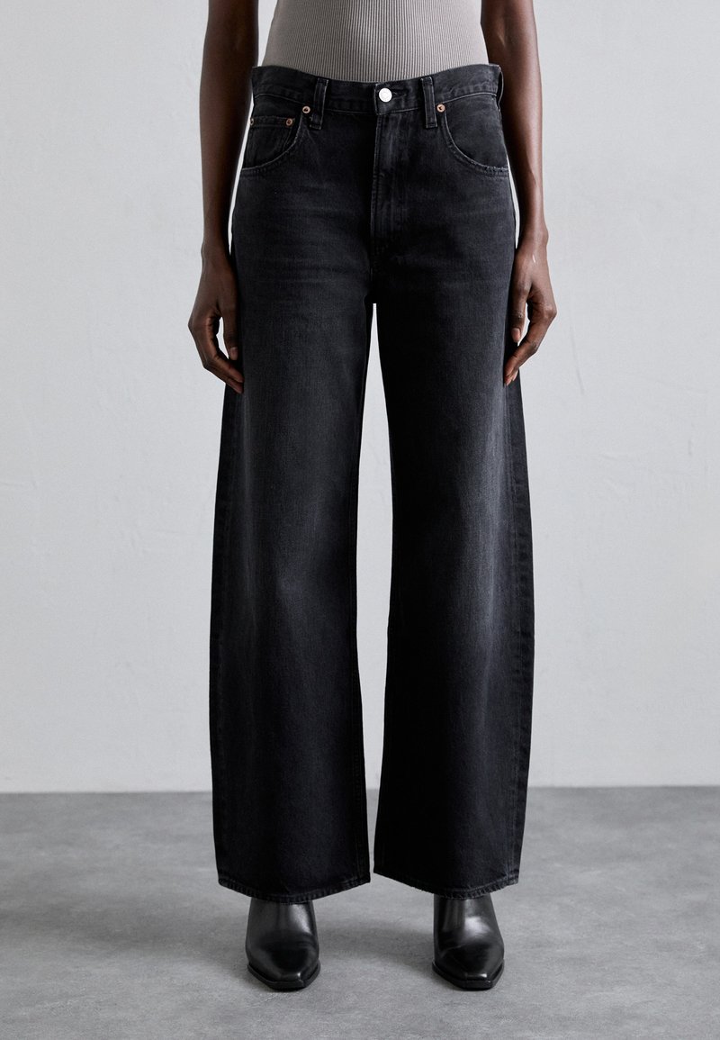 Wide-leg black denim jeans with a high waist, featuring copper button hardware and front pockets, paired with black square-toe boots.