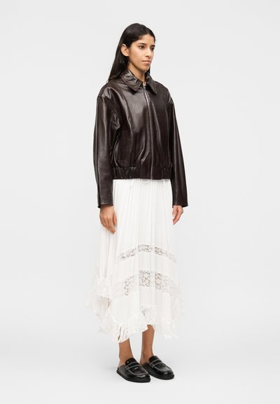 Γυναίκα που φοράει σκούρο καφέ δερμάτινο μπουφάν με φερμουάρ, λευκή πλισέ μίντι φούστα με ενθέσεις δαντέλας και μαύρα μοκασίνια χωρίς κορδόνια, στέκεται πλάγια.