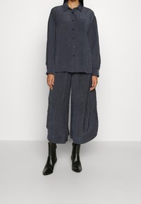 Woman wearing dark gray crinkled fabric button-up shirt with matching wide-leg cropped pants and black ankle boots.