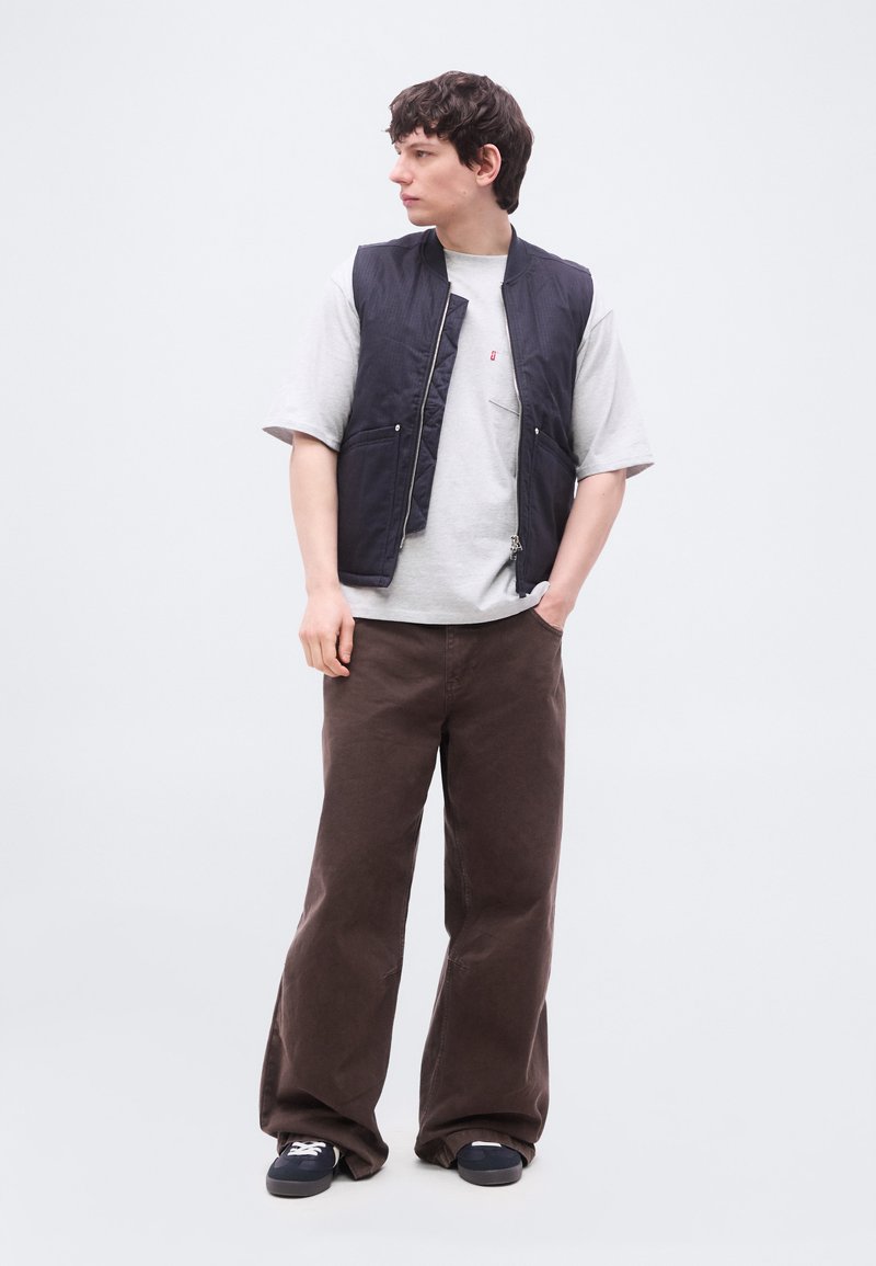 Giovane uomo in piedi con le mani nelle tasche, indossa una T-shirt grigia, un gilet blu scuro, pantaloni larghi marroni e sneakers nere, guarda di lato.