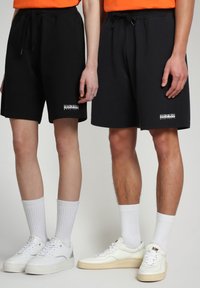 Pantaloni corti in cotone neri con vita elastica e cordino, con un logo bianco sull'orlo, abbinati a sneaker bianche e calzini.