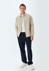 Camisa beige de botones con dos bolsillos en el pecho, combinada con pantalones navy. El modelo lleva zapatillas blancas; el fondo es completamente blanco.
