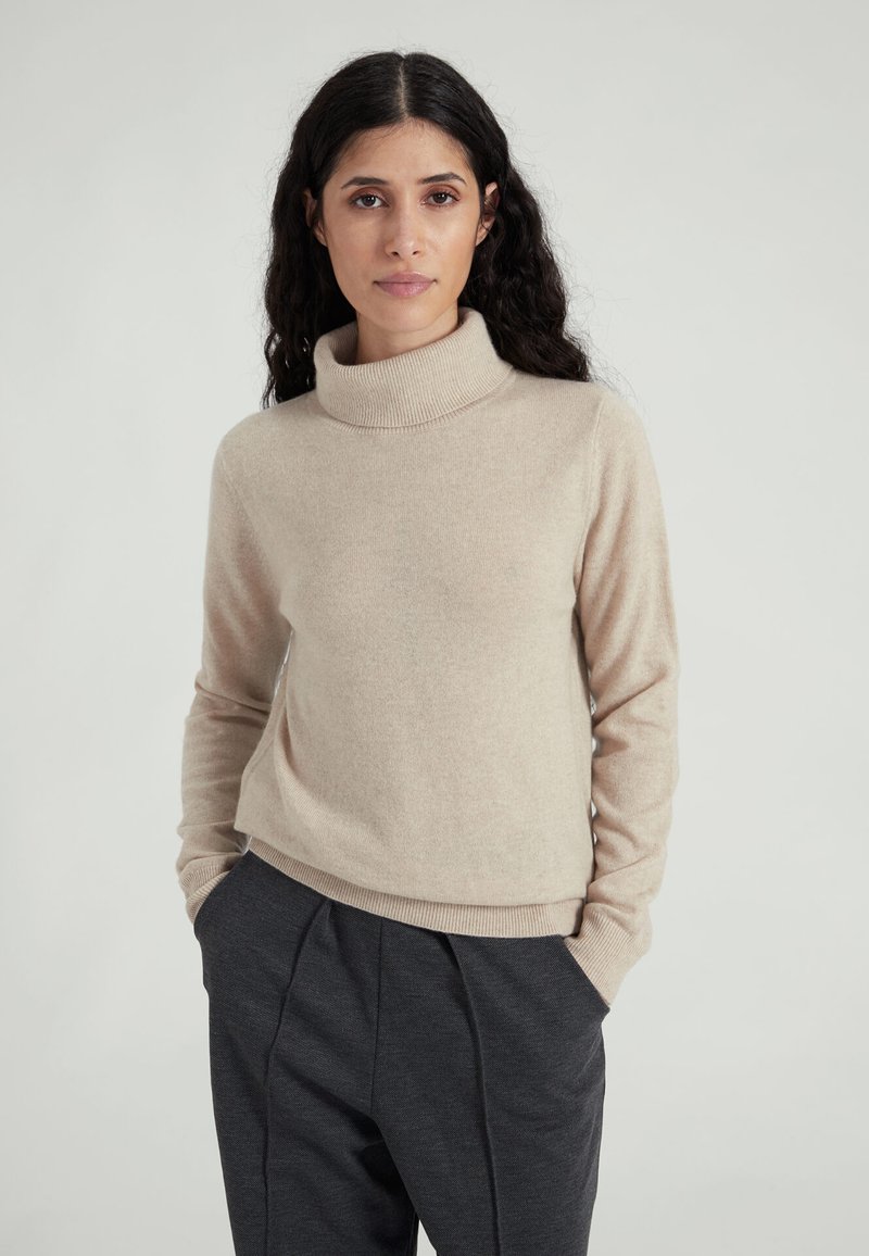 Maglione beige a collo alto in tessuto morbido a maglia con maniche lunghe e orlo a coste, abbinato a pantaloni grigio scuro con tasche laterali.